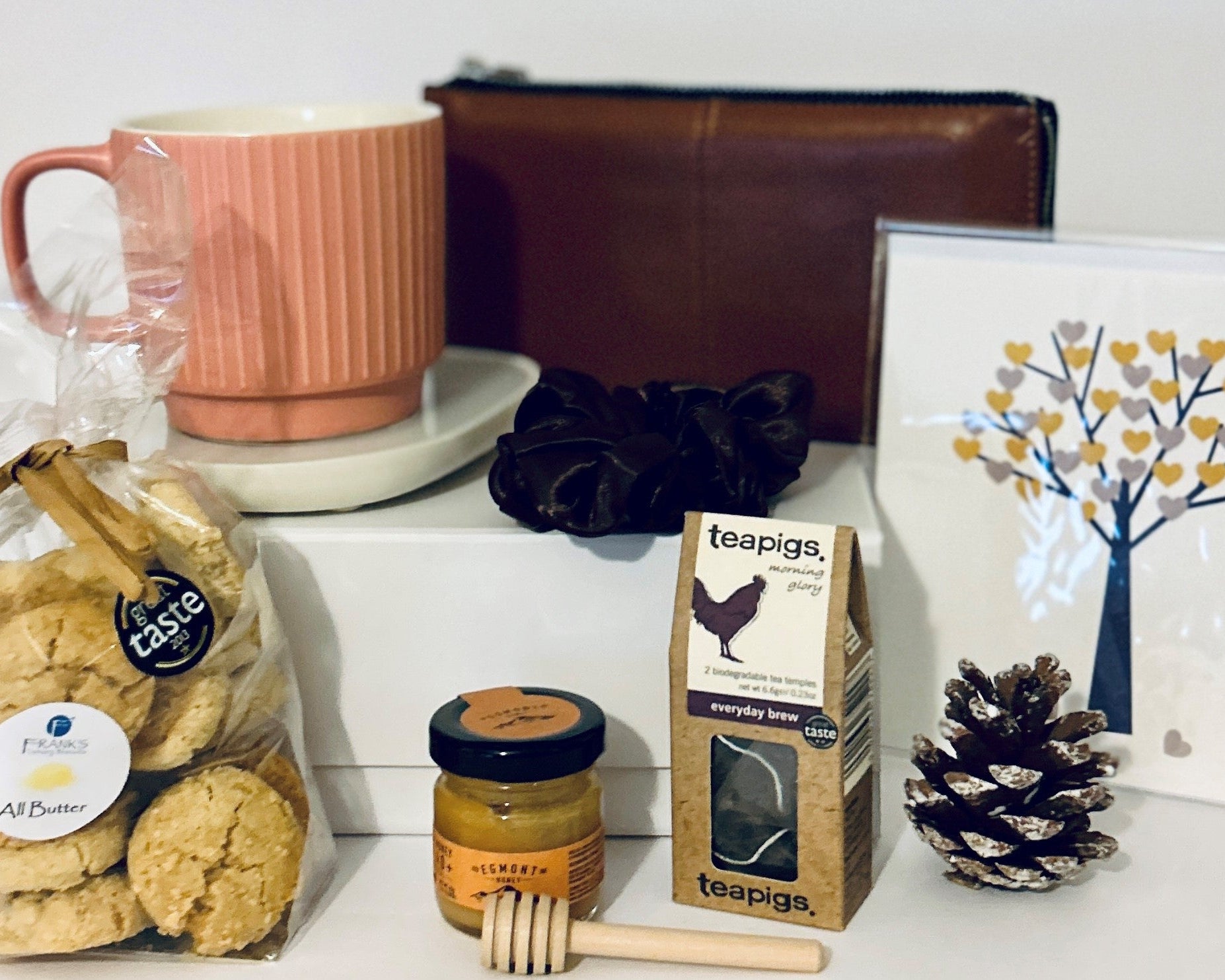 Assorted items including cookies, a mug, a jar of honey, a faux leather purse, a cup warmer, tea, a scrunchie, a cone and a card on a white surface.