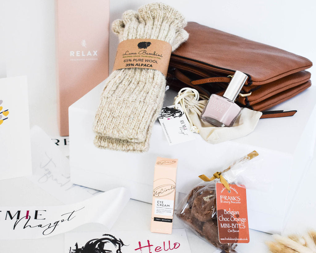 Collection of beauty and lifestyle products, including a brown leather clutch, nail varnish, diffuser, Alpaca socks, biscuits and Upcircle eye cream on a white surface.